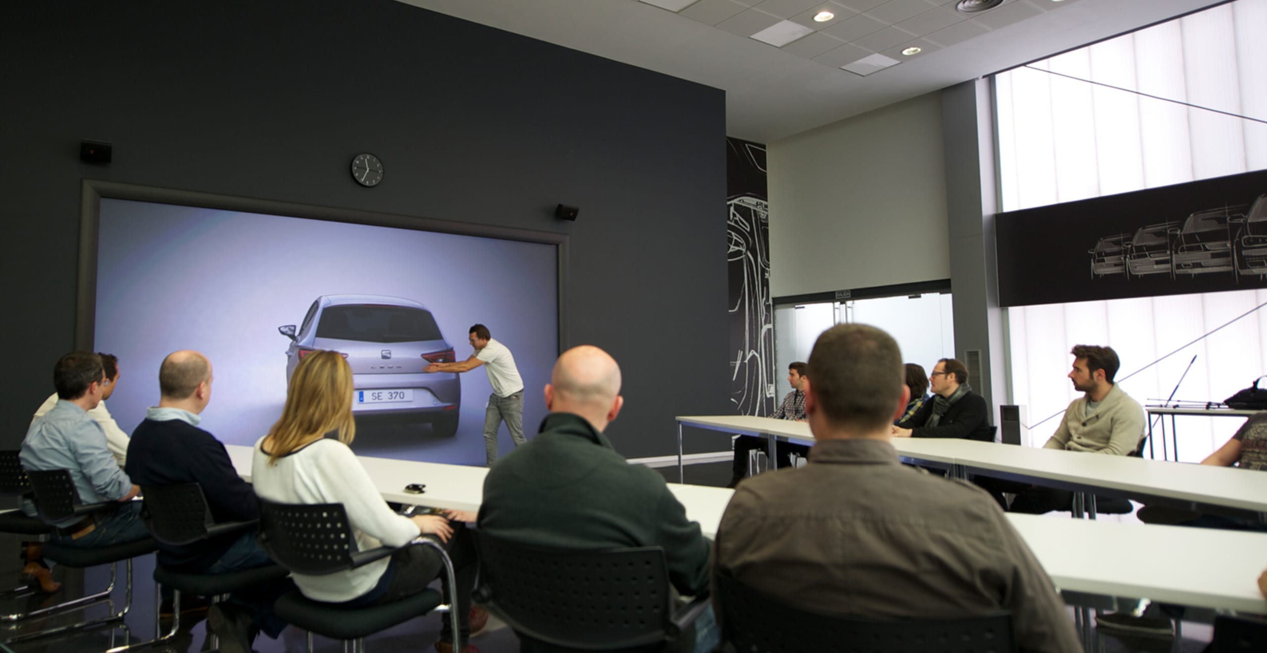 Hombre gesticulando hacia la vista posterior del diseño del auto de León SEAT con personas sentadas en la mesa mirando - SEAT Recursos Humanos