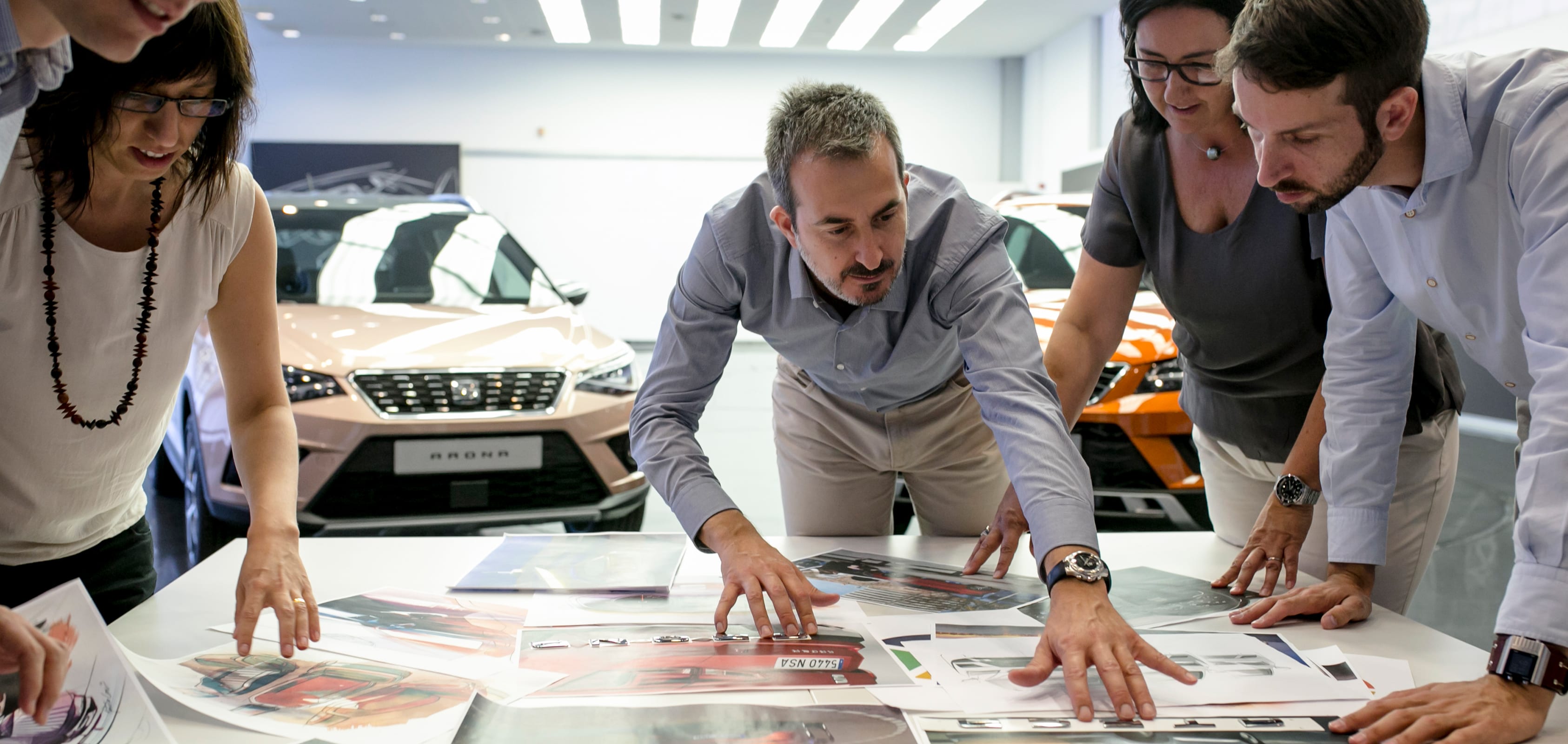 Cuatro personas inspeccionan fotografías de diseños de automóviles SEAT con modelos SEAT Arona en el fondo - Recursos Humanos de SEAT
